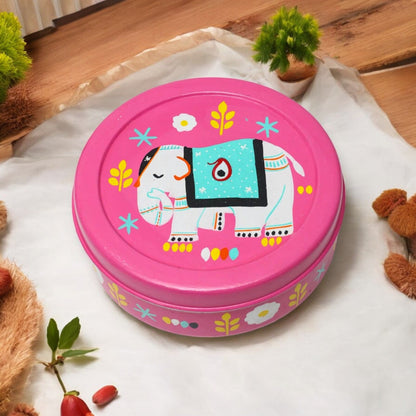 A round, pink spice box with a hand-painted white elephant design and colorful floral motifs. The box is placed on a white surface with a plant and a teddy bear in the background.