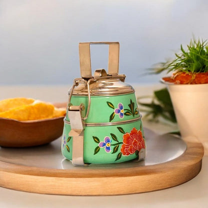 A hand-painted 2-tier steel tiffin box in green with vibrant floral patterns, displayed on a wooden tray.