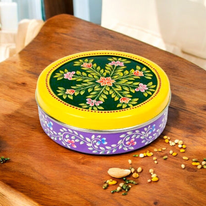A round spice box with a green and purple floral hand-painted design on the lid, placed on a wooden surface with spices scattered around.