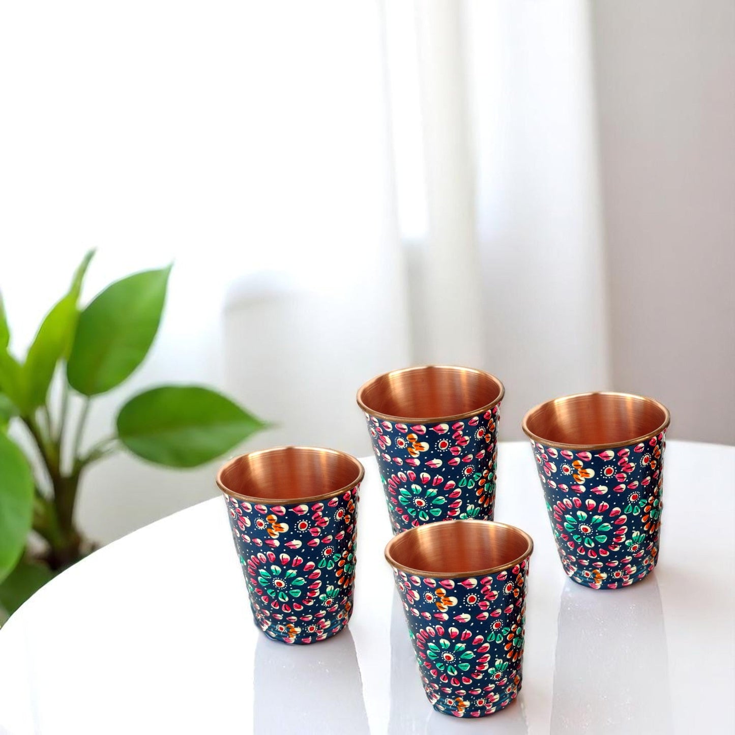 Four copper cups with floral patterns on a white surface, with a plant in the background.