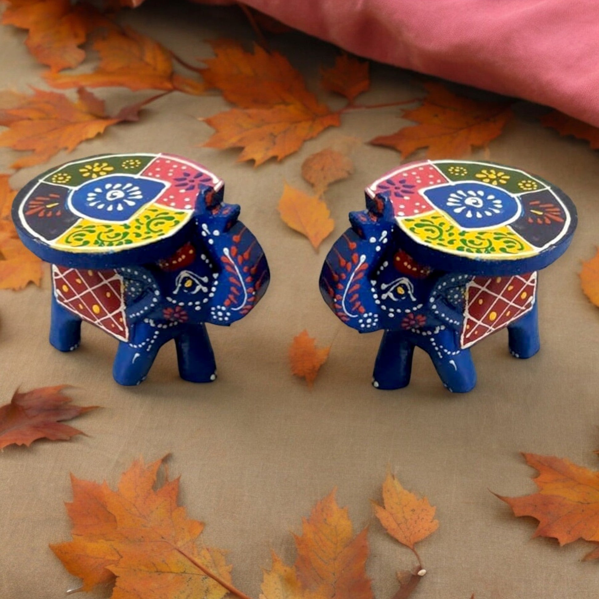 Two hand-painted wooden elephant stools with colorful patterns, one with a turquoise body and red saddle blanket, the other with a blue body and yellow saddle blanket, both with plush, removable cushions on their backs, placed on a surface with autumn leaves around them.