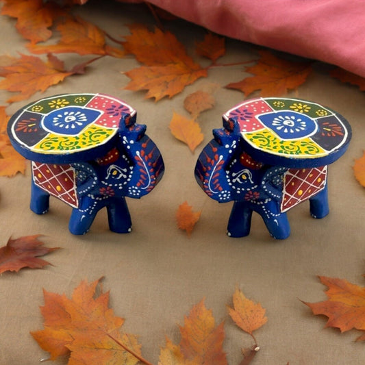 Two hand-painted wooden elephant stools with colorful patterns, one with a turquoise body and red saddle blanket, the other with a blue body and yellow saddle blanket, both with plush, removable cushions on their backs, placed on a surface with autumn leaves around them.