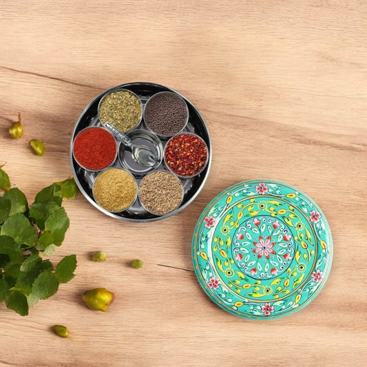 A round, green hand-painted spice box with seven compartments, displayed alongside a small matching spoon and a decorative element on a wooden surface.