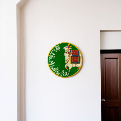 Decorative wall plate with a design of a goat and gift box on a green background, mounted on a white wall.