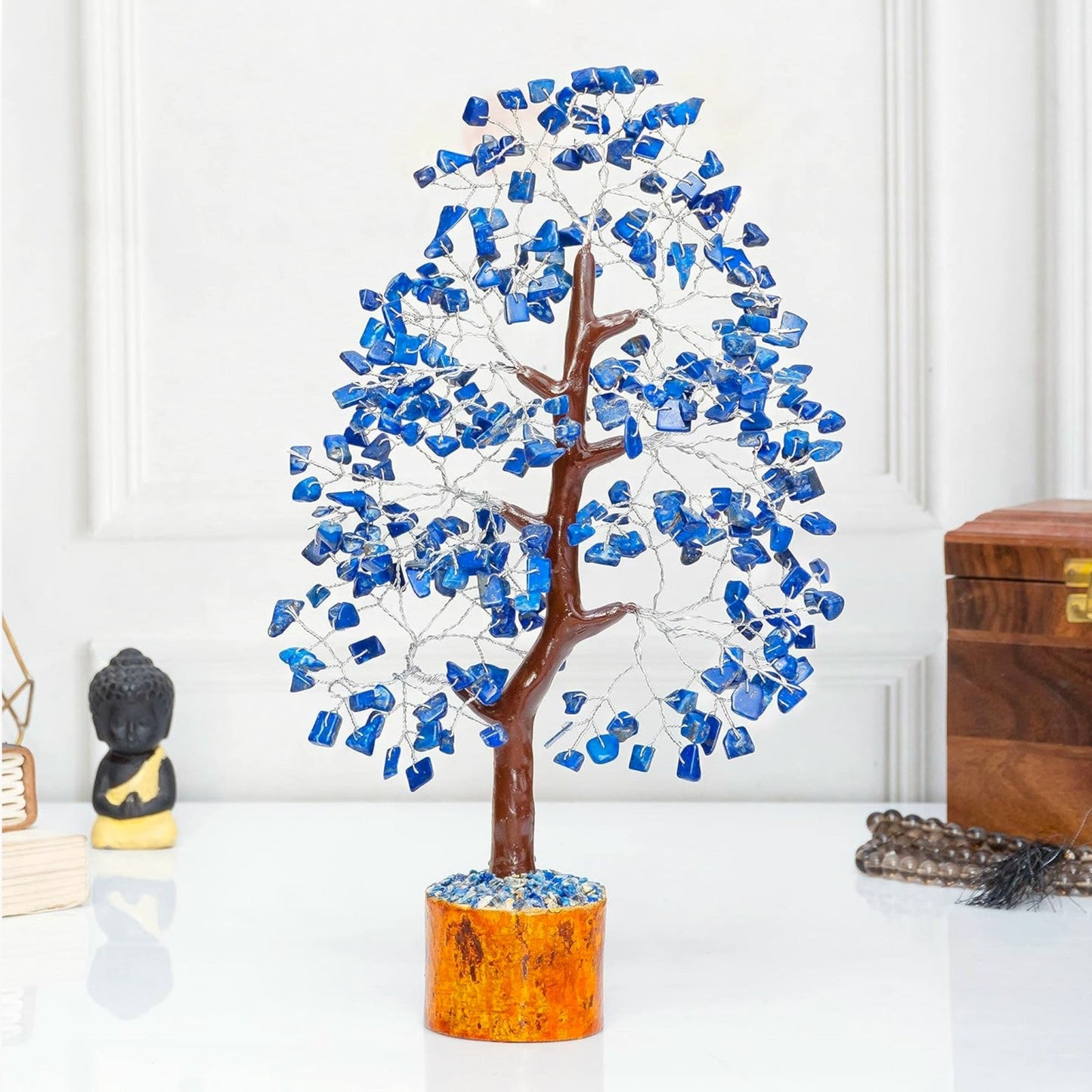 Decorative tree with blue stones on a white surface with a wooden box and small statue in the background.