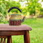 A colorful hand-painted aluminum tea kettle figurine with a wooden handle, displayed on a wooden surface.