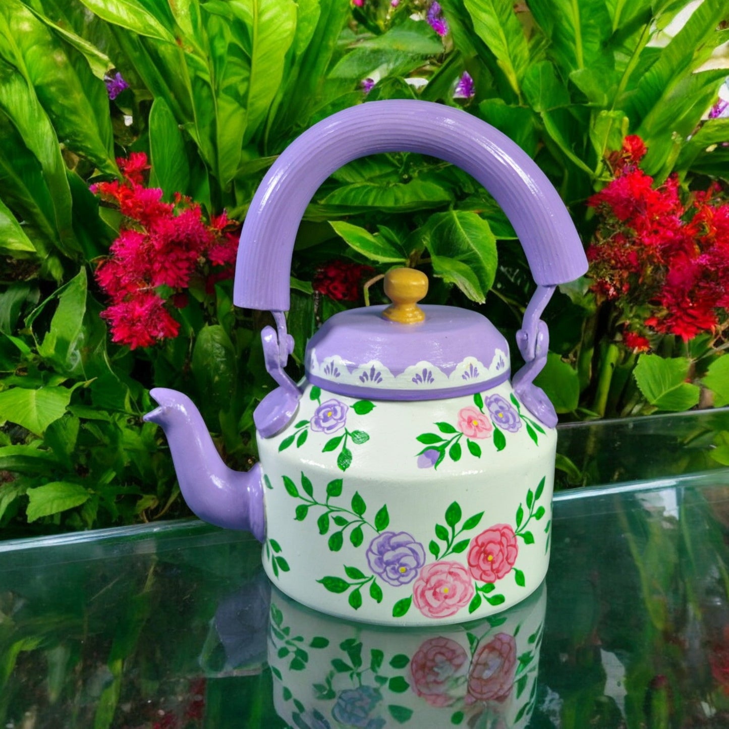 An aluminum tea kettle with a hand-painted floral design in multicolors, with a wooden handle, placed against a backdrop of green plants with red flowers.